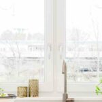 A clean, minimalistic, white and neutral, modern kitchen with a sink, bowl of fruit, and, behind it, a bright window.