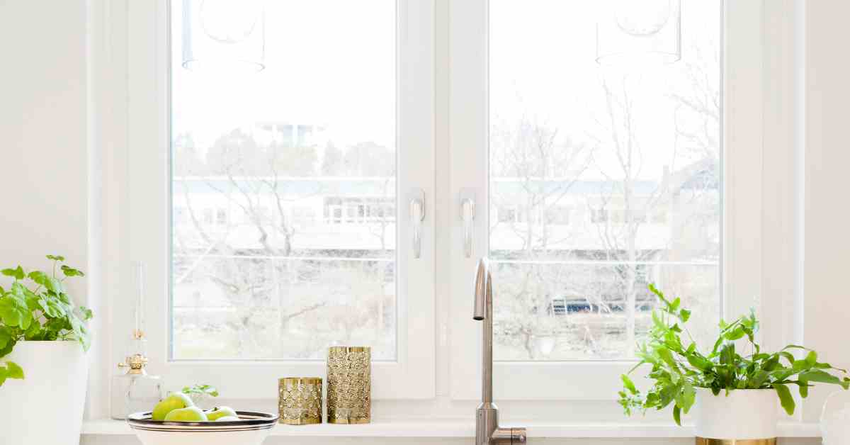 A clean, minimalistic, white and neutral, modern kitchen with a sink, bowl of fruit, and, behind it, a bright window.