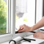 A pair of hands attach black foam to a white winow pane on a sunny day. A tube of caulk sits on the window sill.