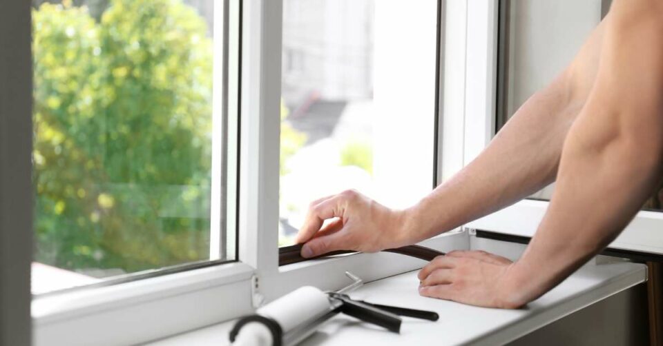 A pair of hands attach black foam to a white winow pane on a sunny day. A tube of caulk sits on the window sill.