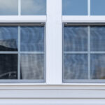 A double-hung vinyl window on a house with white vinyl siding. A blue sky with clouds is visible in the window’s reflection.