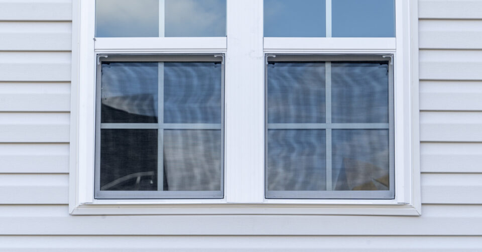 A double-hung vinyl window on a house with white vinyl siding. A blue sky with clouds is visible in the window’s reflection.