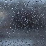 A window pane with raindrops on it. The view outside the window is blurry with abstract gray, brown, and white shapes.