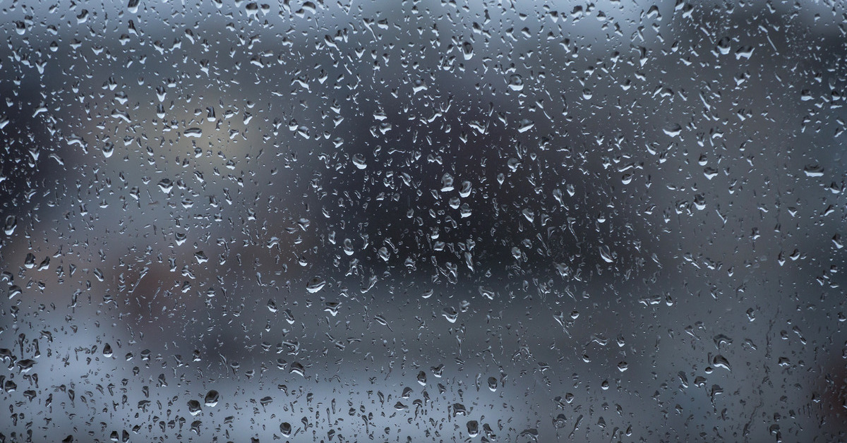 A window pane with raindrops on it. The view outside the window is blurry with abstract gray, brown, and white shapes.