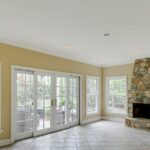 An empty sunroom with tiled floors, dark yellow walls, and a stone fireplace. Metal furniture sit on the patio outside.