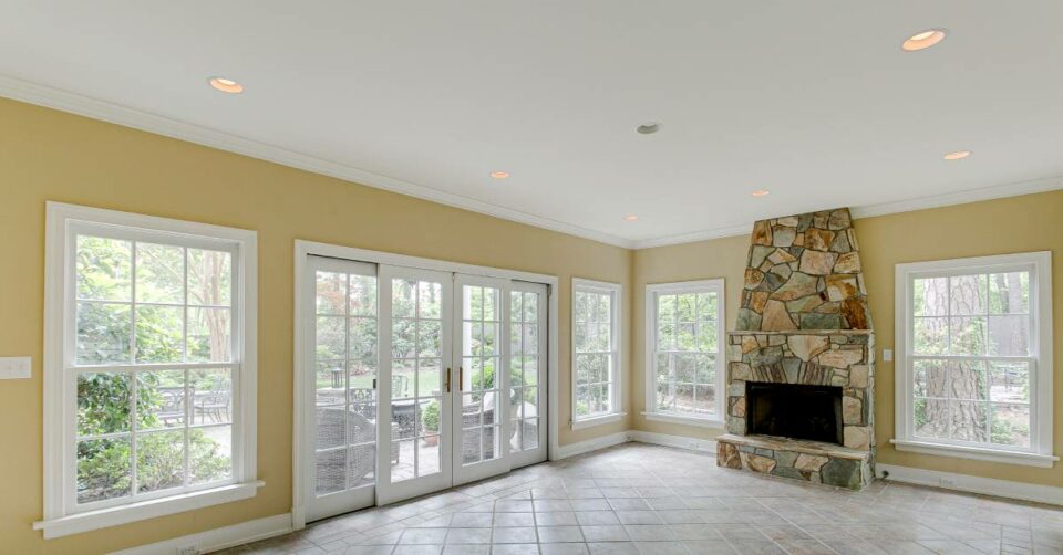 An empty sunroom with tiled floors, dark yellow walls, and a stone fireplace. Metal furniture sit on the patio outside.