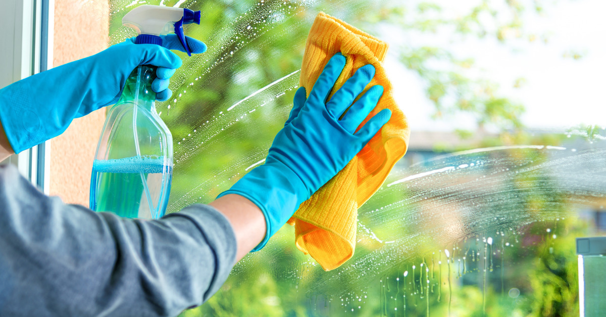 Two gloved hands, one holding a clear spray bottle with blue liquid and one holding a yellow rag, clean a window overlooking trees and other buildings.