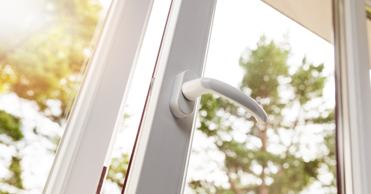 A slightly open white PVC window with a white handle on it. Through the glass a few trees with green leaves are blurry but visible. It is sunny.