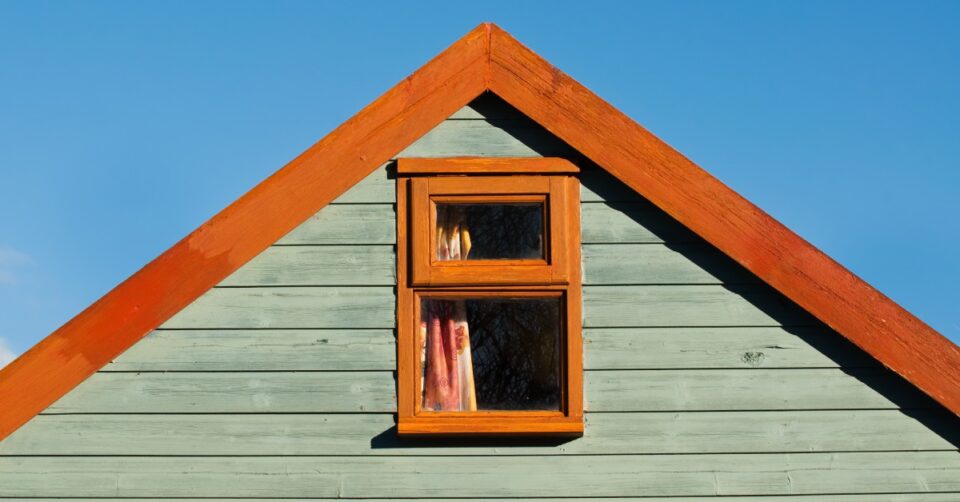 The top of a building with green siding and a wood-clad window and roof. The sky behind the structure is blue.