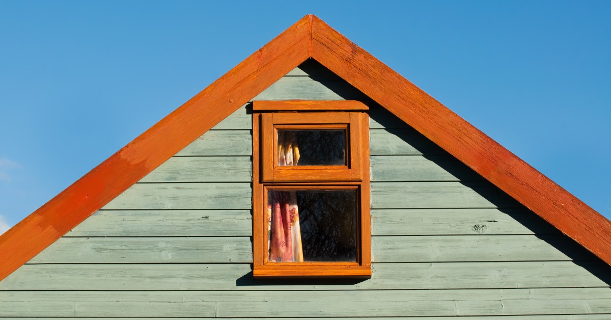 The top of a building with green siding and a wood-clad window and roof. The sky behind the structure is blue.