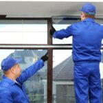 A man in a blue jumpsuit and hat points to where another man in the same attire is installing a window.