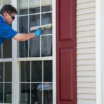 A man in a blue shirt and sunglasses standing on a ladder leans over to use a soapy squeegee on a window.