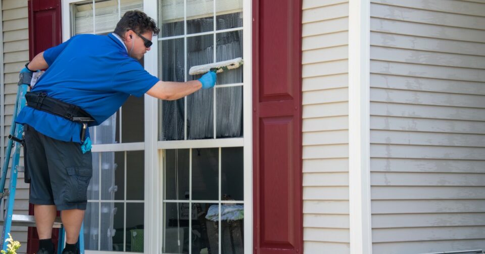 A man in a blue shirt and sunglasses standing on a ladder leans over to use a soapy squeegee on a window.