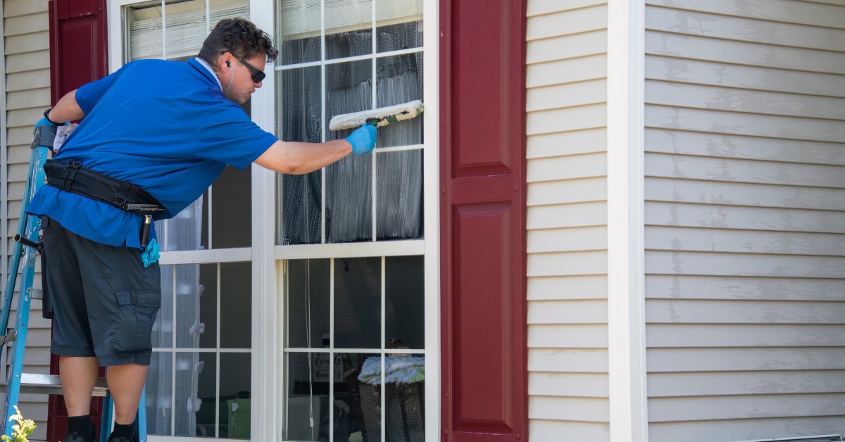 A man in a blue shirt and sunglasses standing on a ladder leans over to use a soapy squeegee on a window.