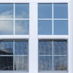 A white vinyl window on the side of a house with white siding. The sky and a cloud are reflected in the window.