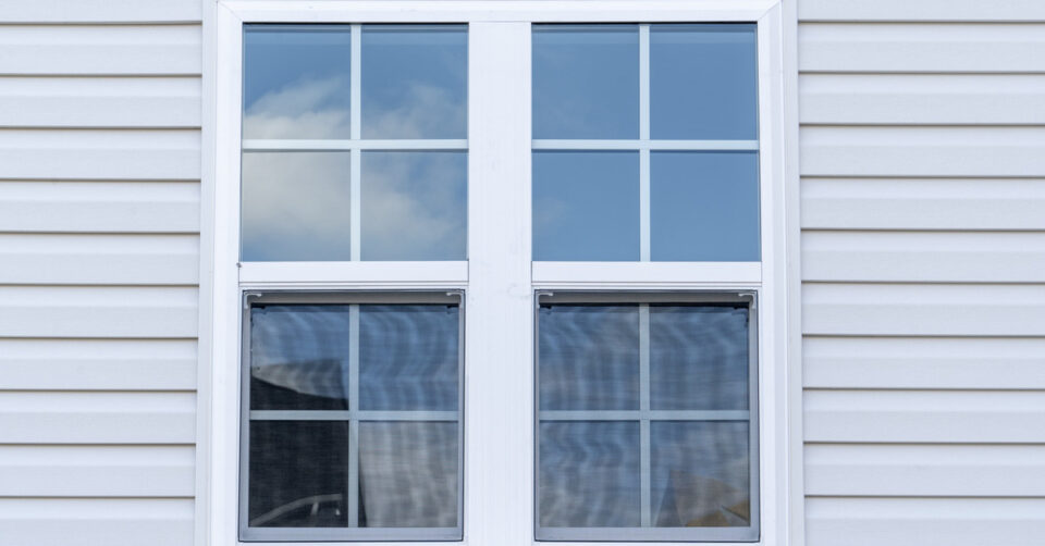 A white vinyl window on the side of a house with white siding. The sky and a cloud are reflected in the window.