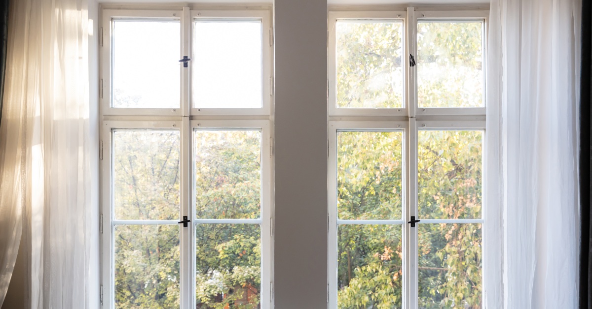 Two closed windows in a room with gray walls. The curtains are open and trees are visible through the windows.