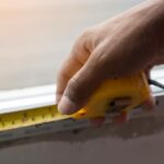 A closeup of a person's hand as they hold measuring tape to what appears to be the bottom of a windowsill.