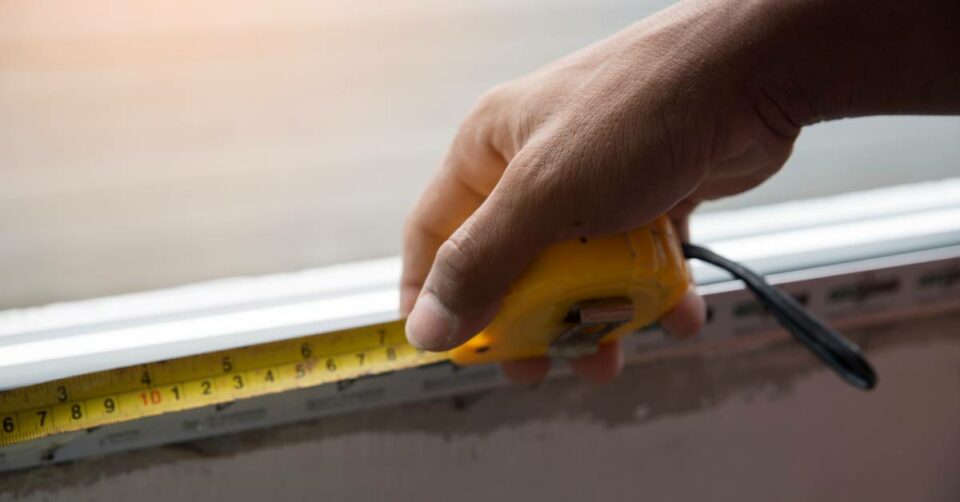 A closeup of a person's hand as they hold measuring tape to what appears to be the bottom of a windowsill.