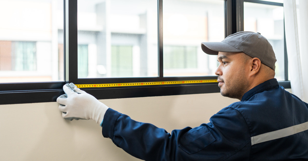 A man wearing a uniform, hat, and gloves holds yellow measuring tape against a window with black trim.