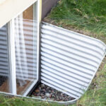 The closeup view of an aluminum window well attached to a basement egress window, surrounded by green grass.