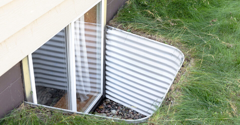 The closeup view of an aluminum window well attached to a basement egress window, surrounded by green grass.