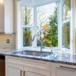 The image of a kitchen design that includes a garden window, white cabinets and drawers, and marble countertops.