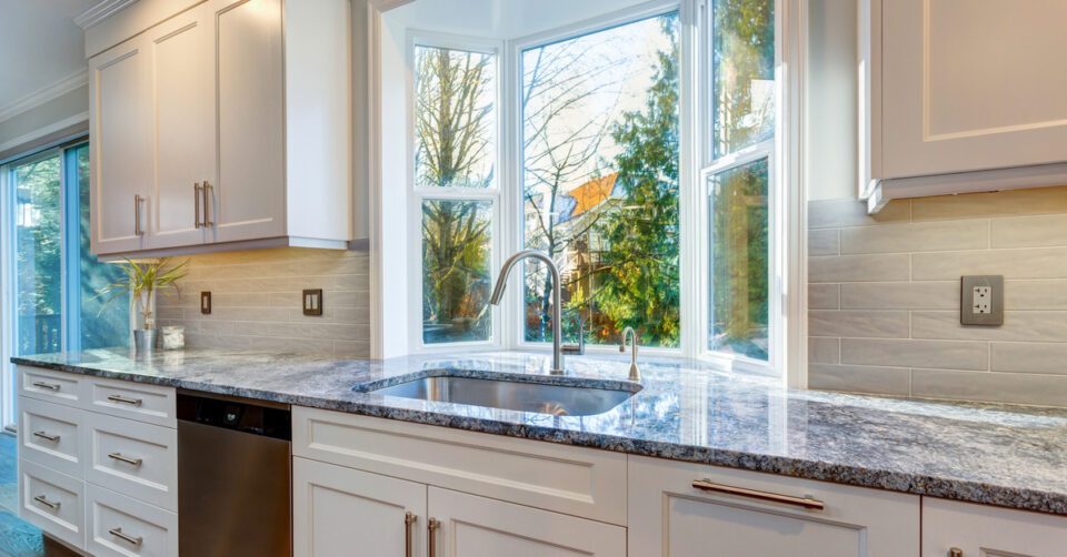 The image of a kitchen design that includes a garden window, white cabinets and drawers, and marble countertops.